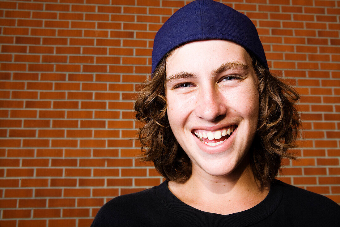 High school aged skateboarder smiling on a brick backdrop, Oceanside, California.
