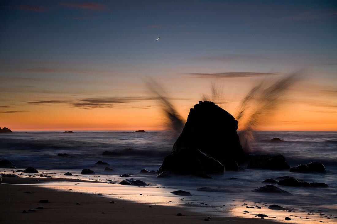 GARRAPATA STATE PARK, CA - DECEMBER 2007: A wave crashes over a large rock as a crescent moon sets over the Pacific along California's Big Sur coast.
