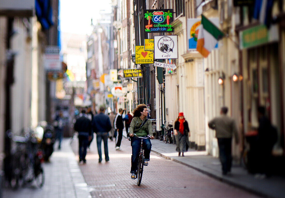 A young woman listening to music on headphones cycles along a lively street  in Amsterdam, The Netherlands Holland,.