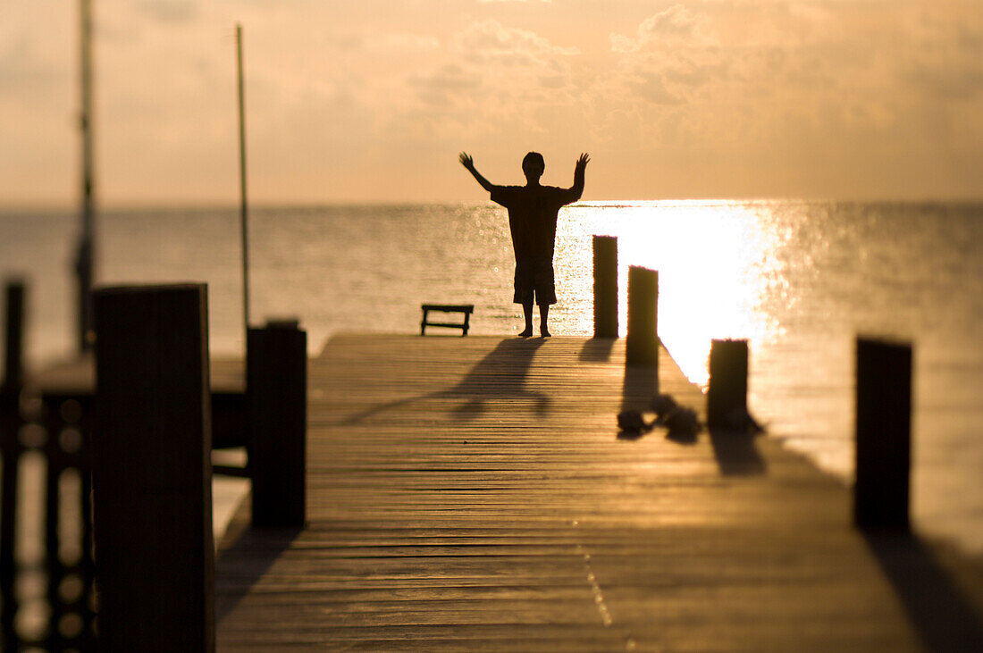 Nico Steele, age 12, holds his arms high while silhouetted against the morning sun on  a wooden dock on Ambergris Caye, Belize.