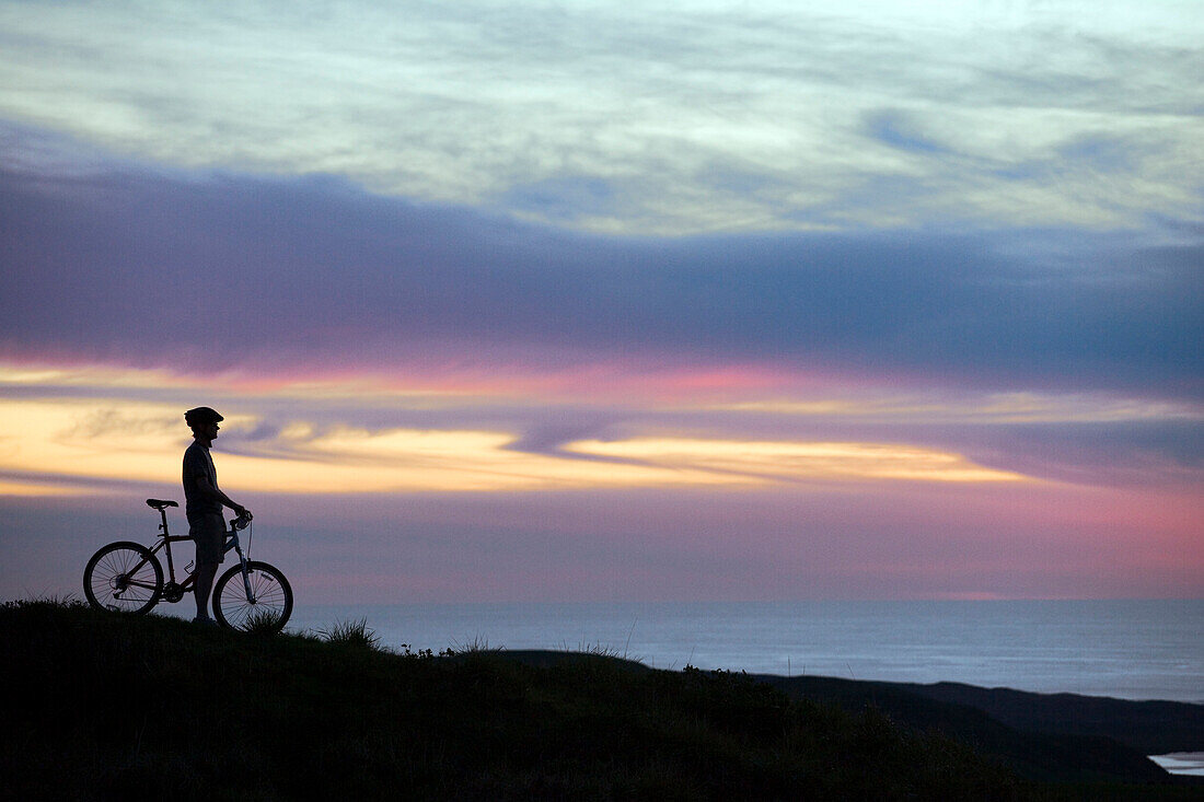 A mountain biker pauses for a moment to enjoy a sunset over the Pacific Ocean.