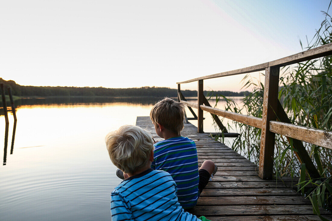 Two boys sitting on a jetty at a lake in dusk, Schorfheide-Chorin Biosphere Reserve, Neudorf, Friedenfelde, Uckermark, Brandenburg, Germany