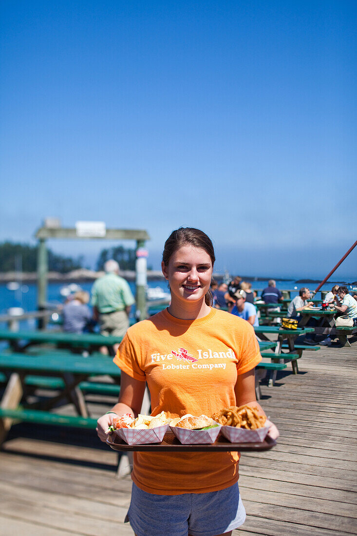 MAINE, USA. A server holds a plate of seafood on a dock on a sunny afternoon.