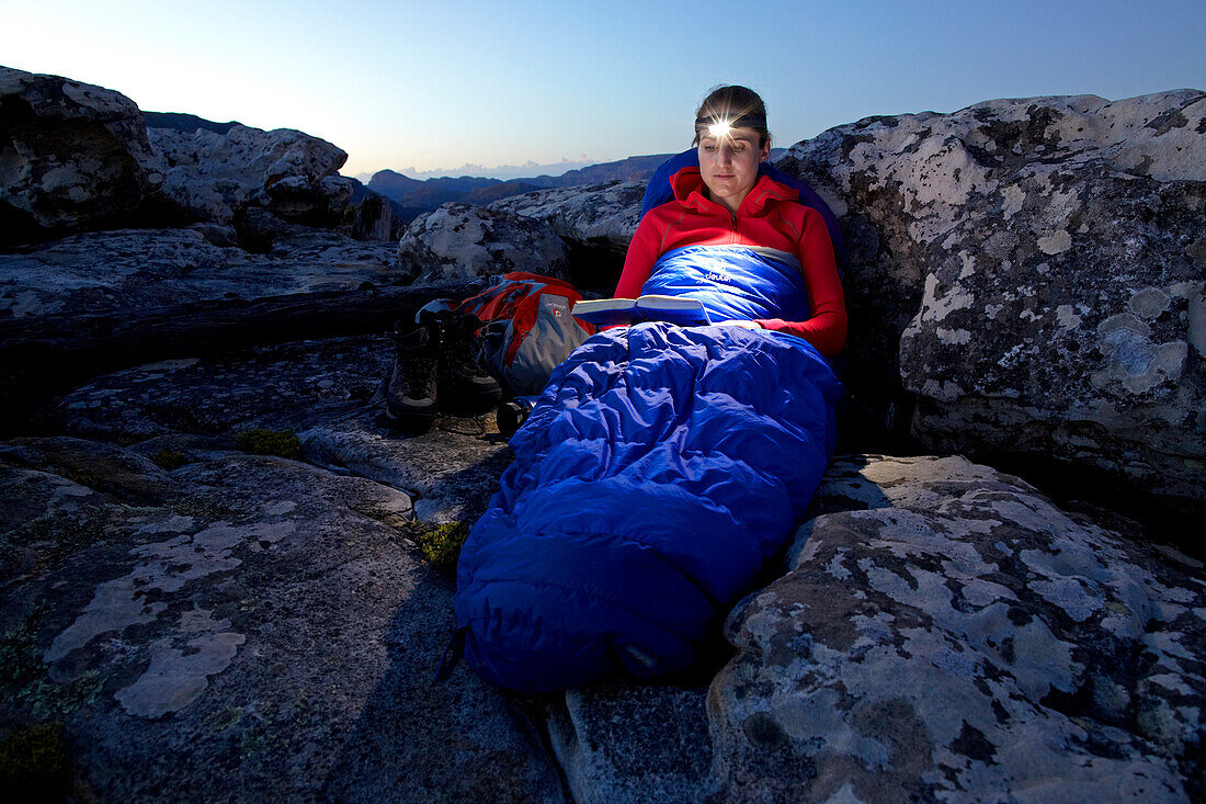Susann Scheller is sitting against a rock in her sleeping bag while reading a book with a headlamp. Table Mountain National Park. South Africa.