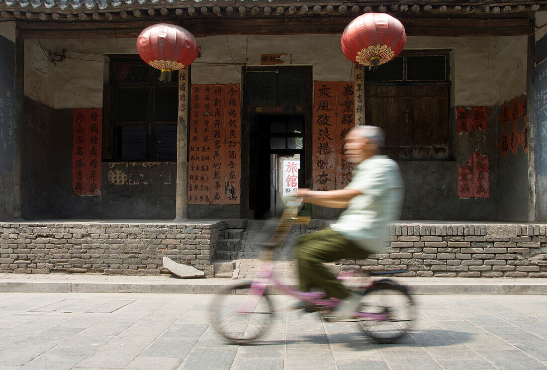 Old Chinese man riding a bike in front of a traditional old street building of Pingyao. Shaanxi Province. China