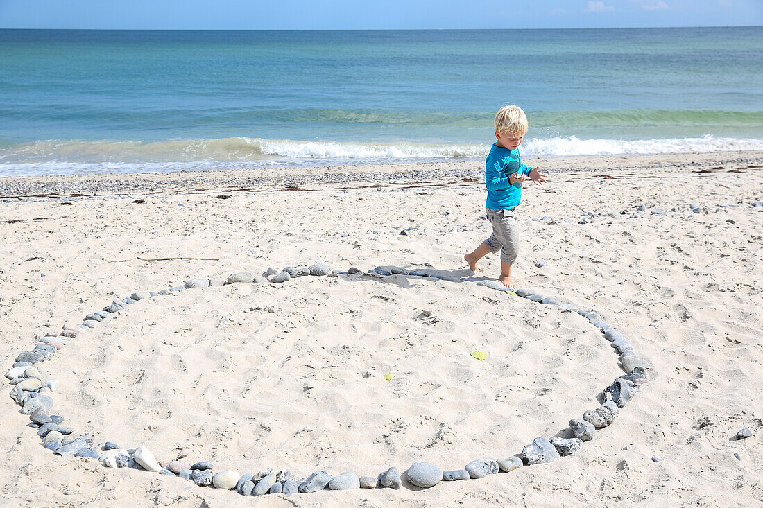 Junge (4 Jahre) umrundet einen Kreis aus Steinen am Strand, Rytsebaek, Insel Mön, Dänemark