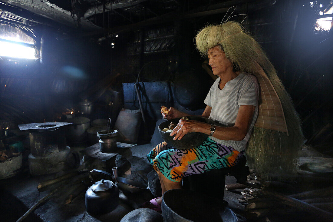 Ivatan woman cooking inside an old kitchen in Chavayan village, wearing a typical straw wig, Sabtang Island, Batanes, Philippines, Asia