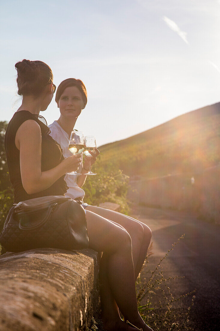 Weinfest an einem lauen Sommerabend: Zwei hübsche junge Frauen mit Weingläsern sitzen auf einer Mauer oberhalb von Weingut am Stein, Würzburg, Franken, Bayern, Deutschland