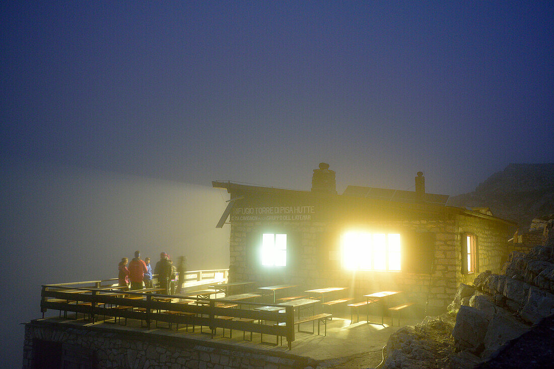 Mehrere Personen stehen vor beleuchteter Latemar-Hütte, Rifugio Torre di Pisa, Latemar, Dolomiten, UNESCO Welterbe Dolomiten, Trentino, Italien