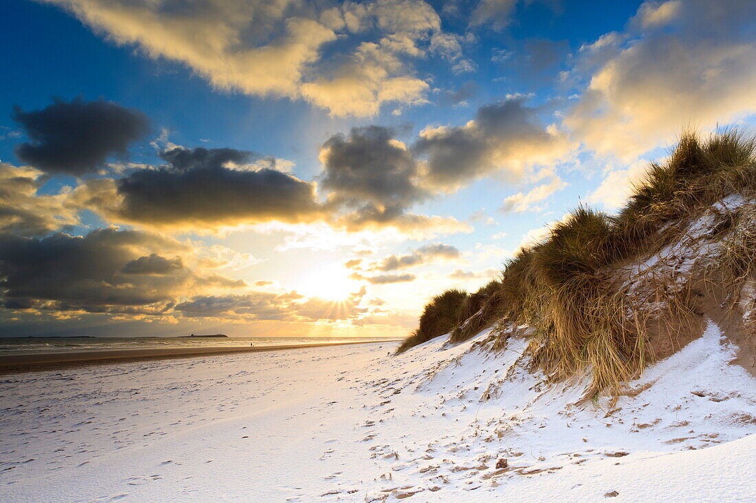 Man walks dog on snow covered Bamburgh beach at dawn with view to Farne Islands, Northumberland, England