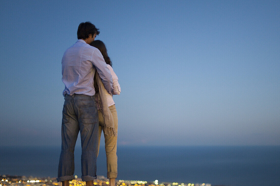 Couple standing on edge of infinity pool at dusk, looking at sea