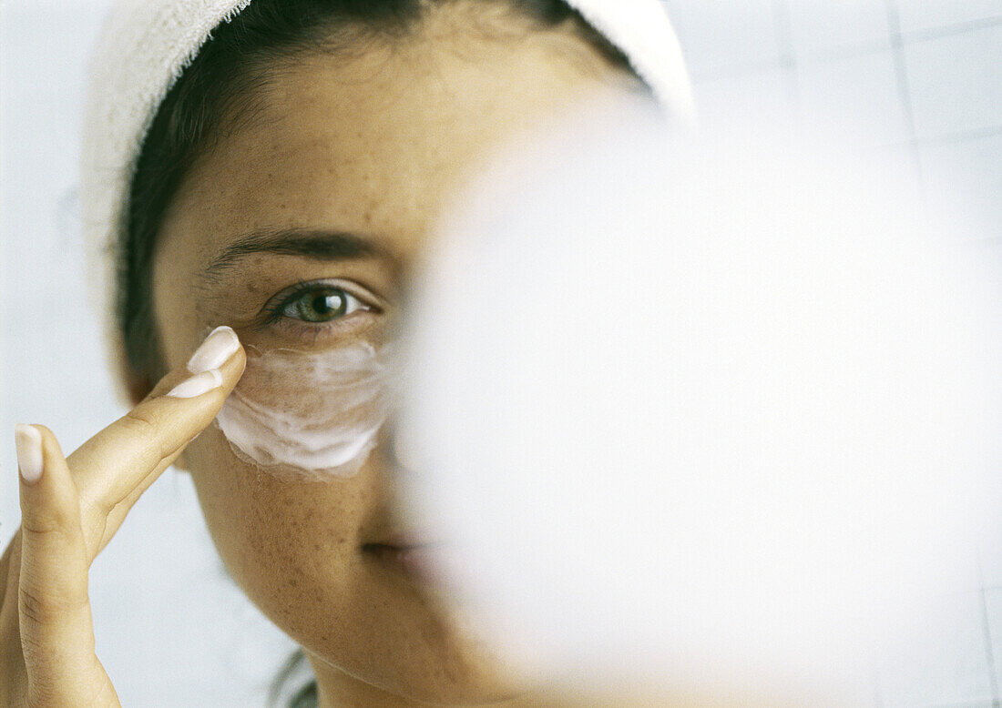 Woman applying moisturizer beneath eye, blurred compact in foreground
