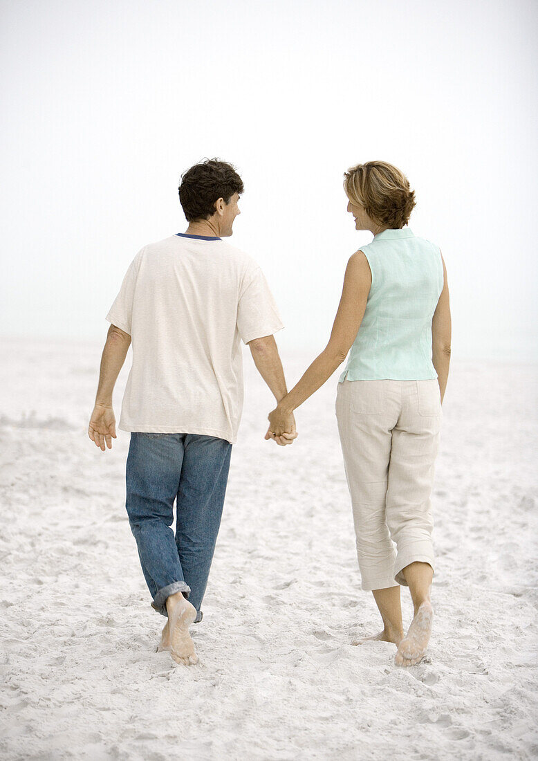 Mature couple walking on beach, holding hands, rear view