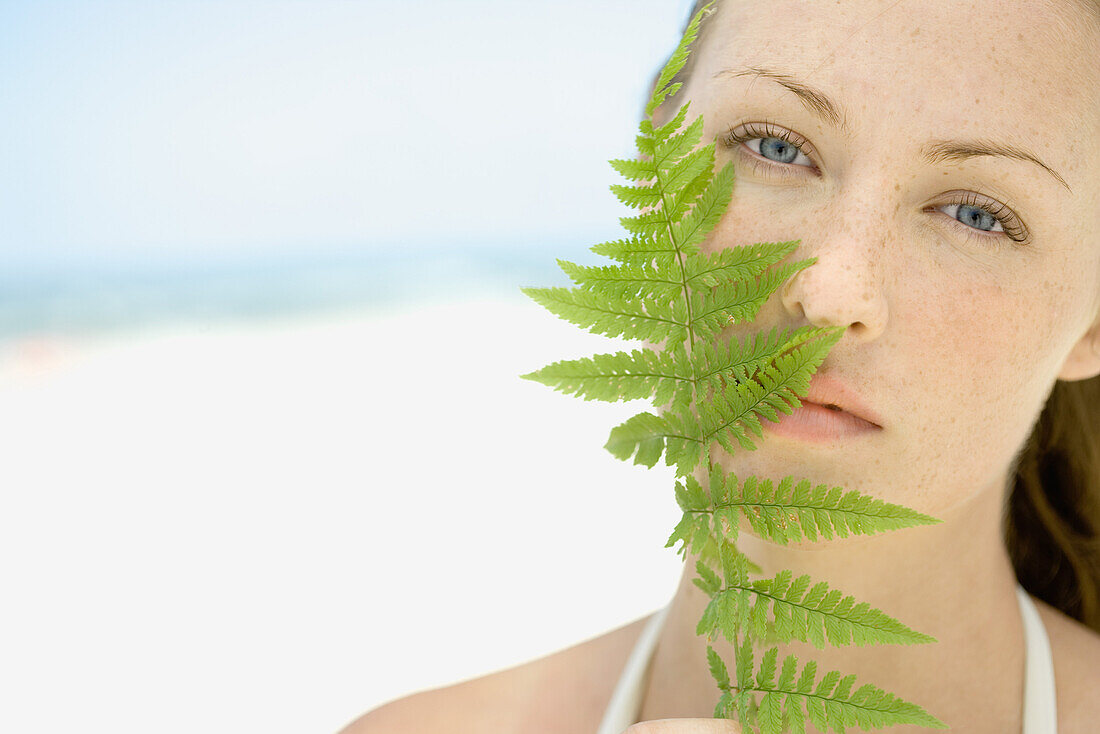 Woman holding fern frond up to face, beach in background