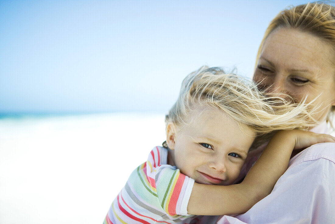 Woman holding daughter on beach, girl smiling at camera as wind tousles her hair