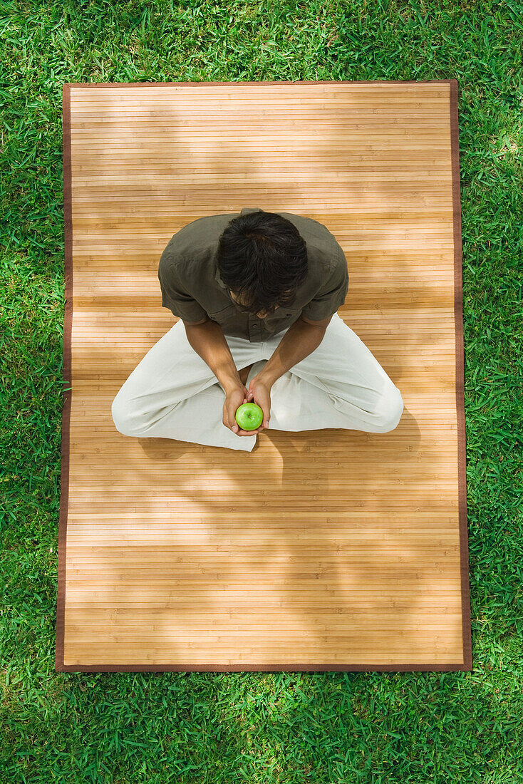 Man sitting crosslegged on mat outdoors, holding apple, high angle view