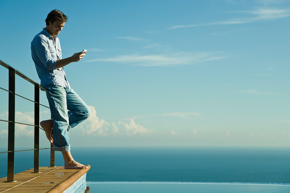 Man standing beside infinity pool, text messaging with cell phone
