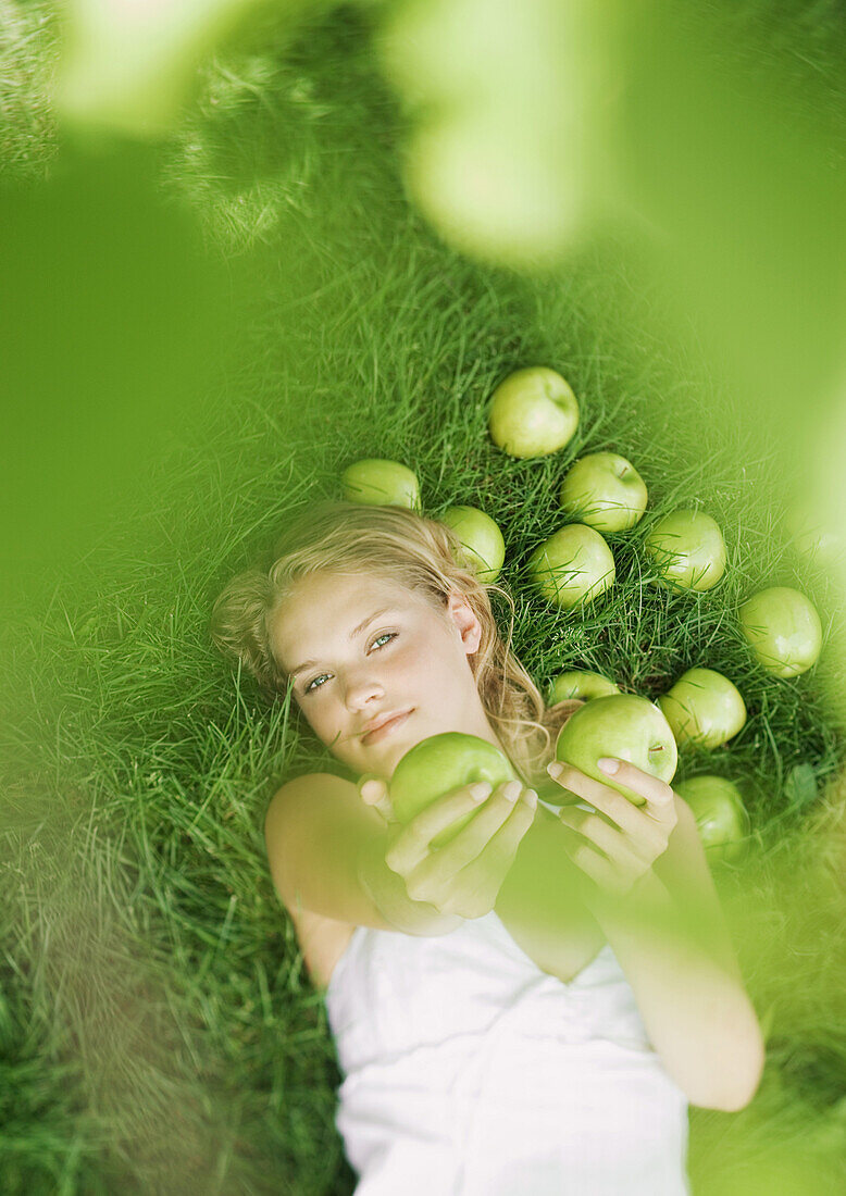 Woman lying in grass, holding up apples toward camera
