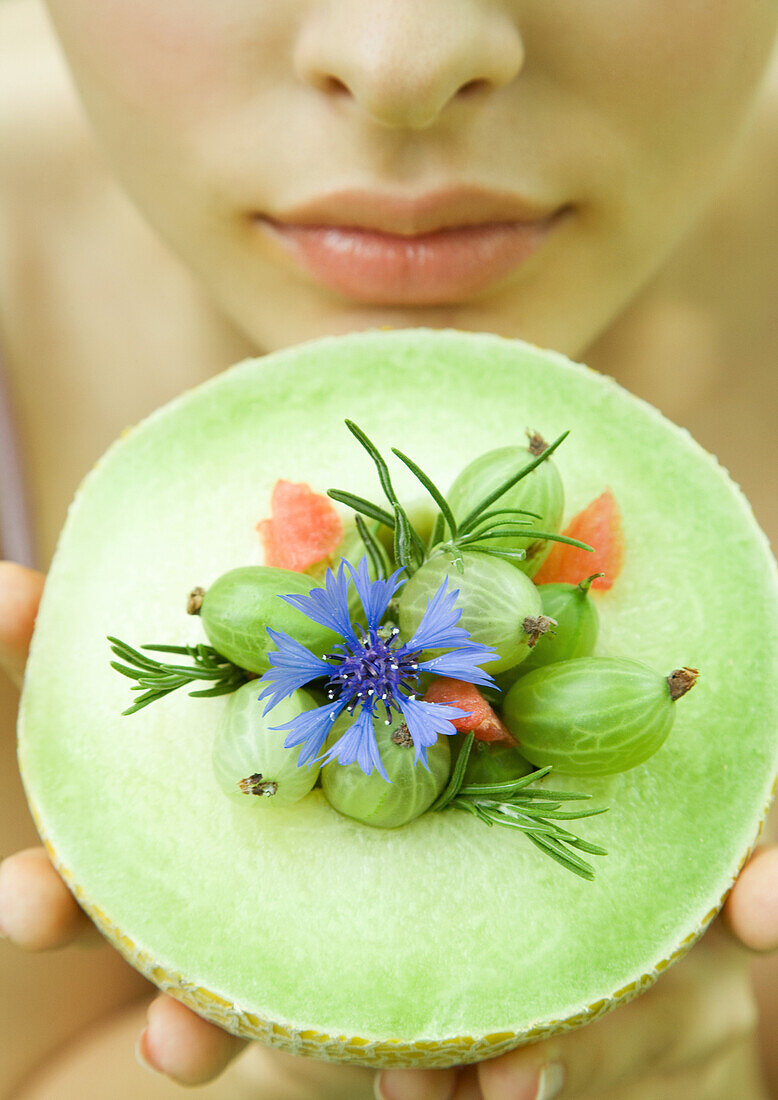 Woman holding up honeydew melon topped with berries, flower and rosemary sprigs, close-up