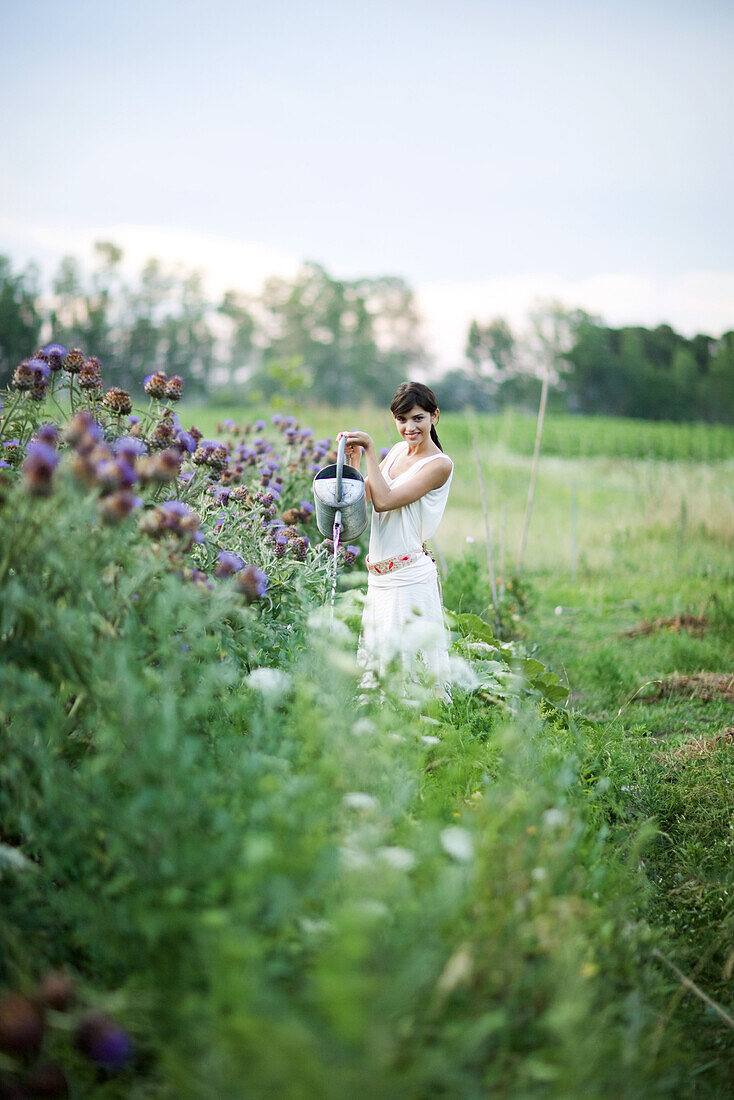 Young woman watering plants with watering can, in mid distance, smiling at camera