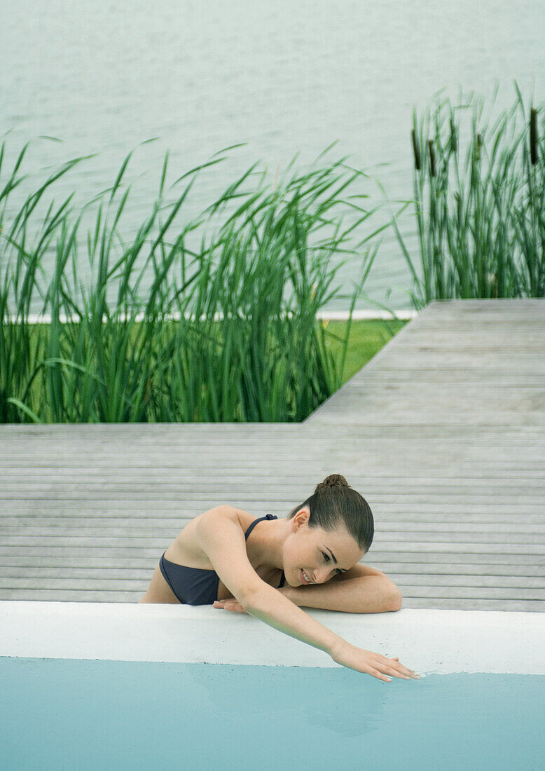 Woman leaning against edge of pool, touching surface of water, lake in background