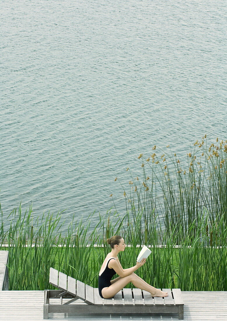 Woman sitting up on lounge chair, reading, next to lake
