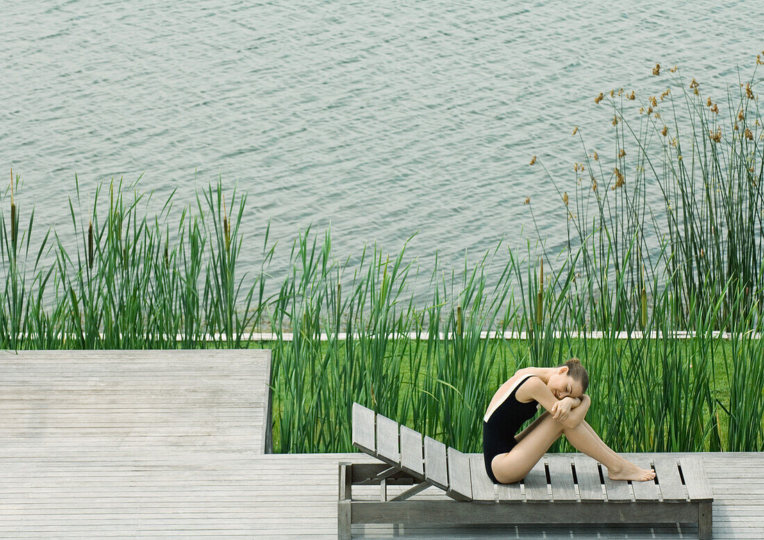 Woman sitting on lounge chair next to lake, hugging knees, resting head on arms