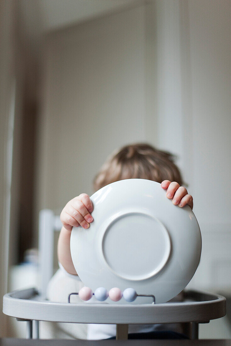 Toddler sitting in high chair, holding plate in front of face