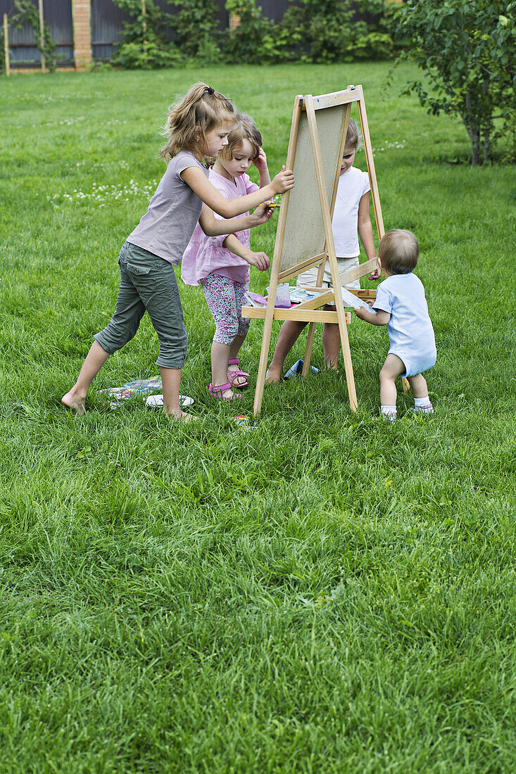 Three young girls and a toddler painting outdoors on an easel