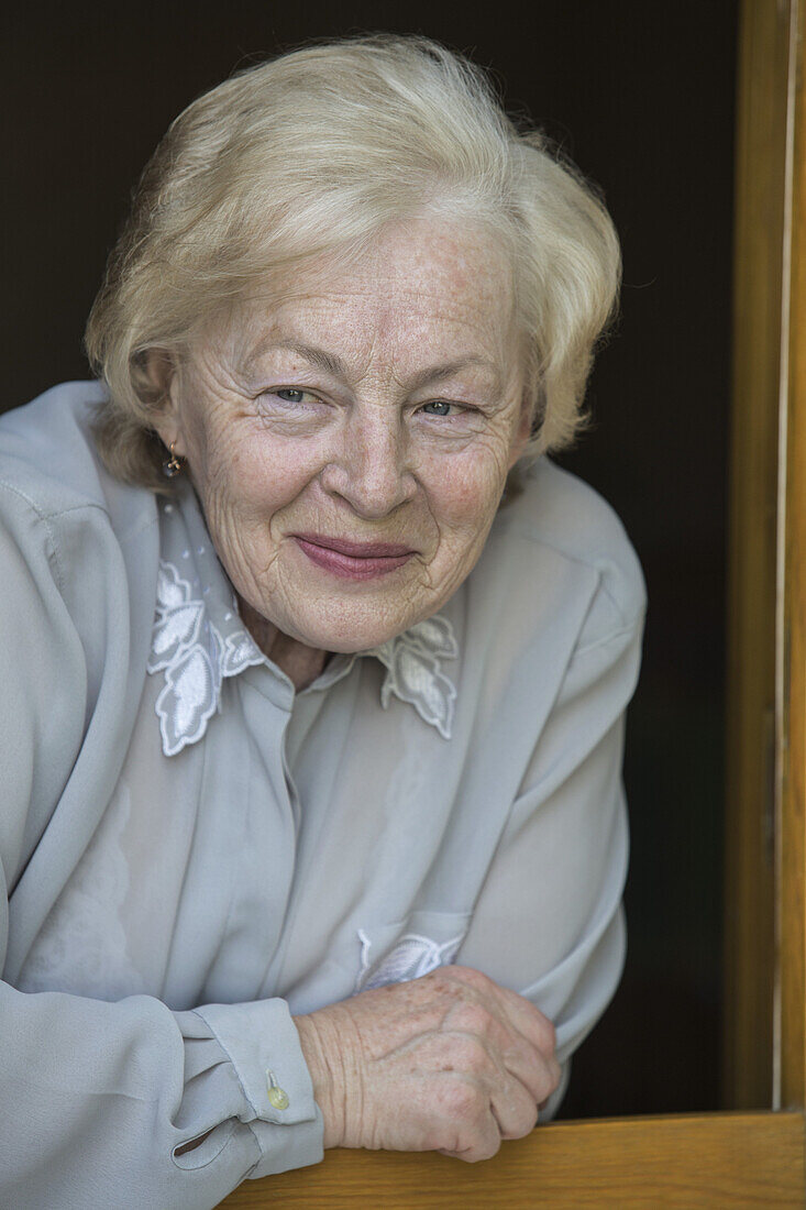 A senior woman leaning on a window sill, smiling while looking away