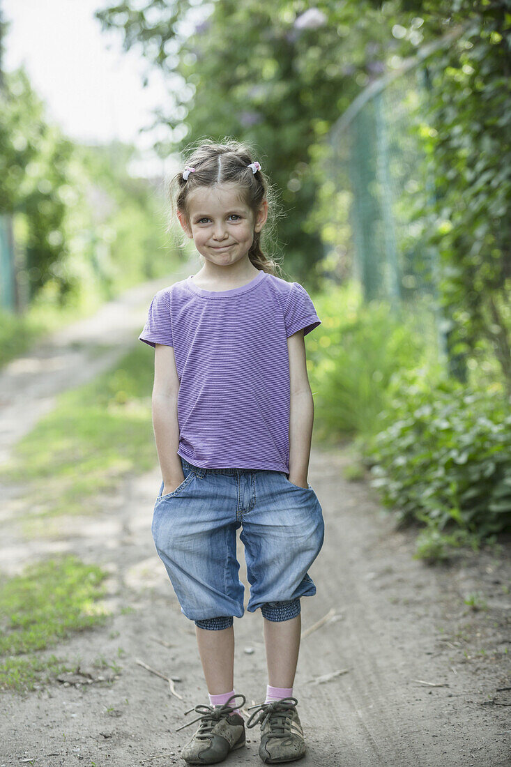A young smiling girl with hands in her pockets, standing on a dirt road