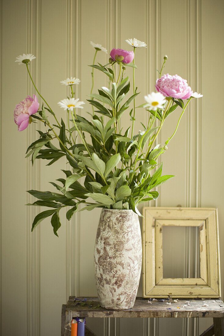 Eine Vase mit Pfingstrosen und Gänseblümchen und ein leerer Bilderrahmen