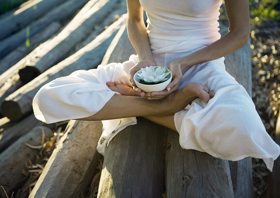 Frau sitzt auf einem Stapel Holzscheite und hält eine Schale mit einer Lotusblume, Brust nach unten