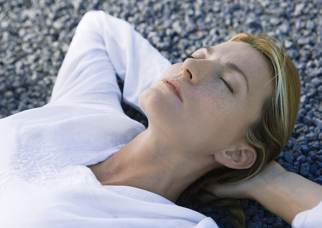 Woman lying on gravel with hands behind head, eyes closed