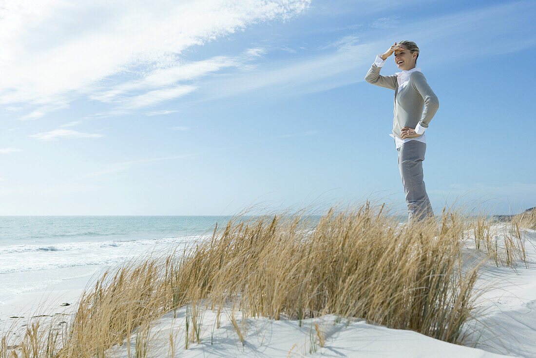 Woman standing on dune, shading eyes and looking into distance, smiling, full length