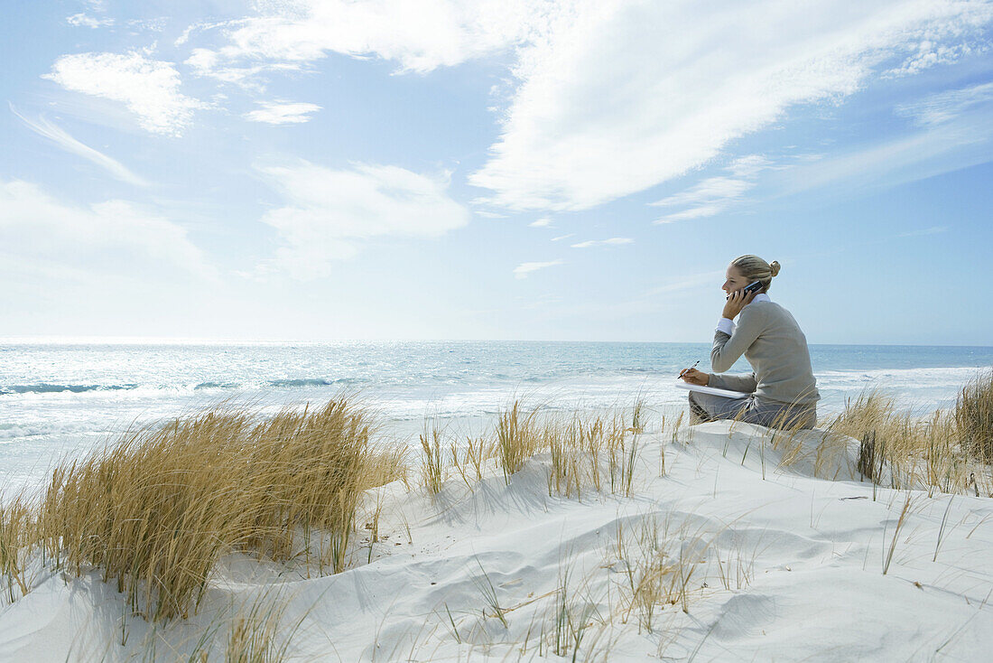 Woman sitting on dunes at beach, using cell phone