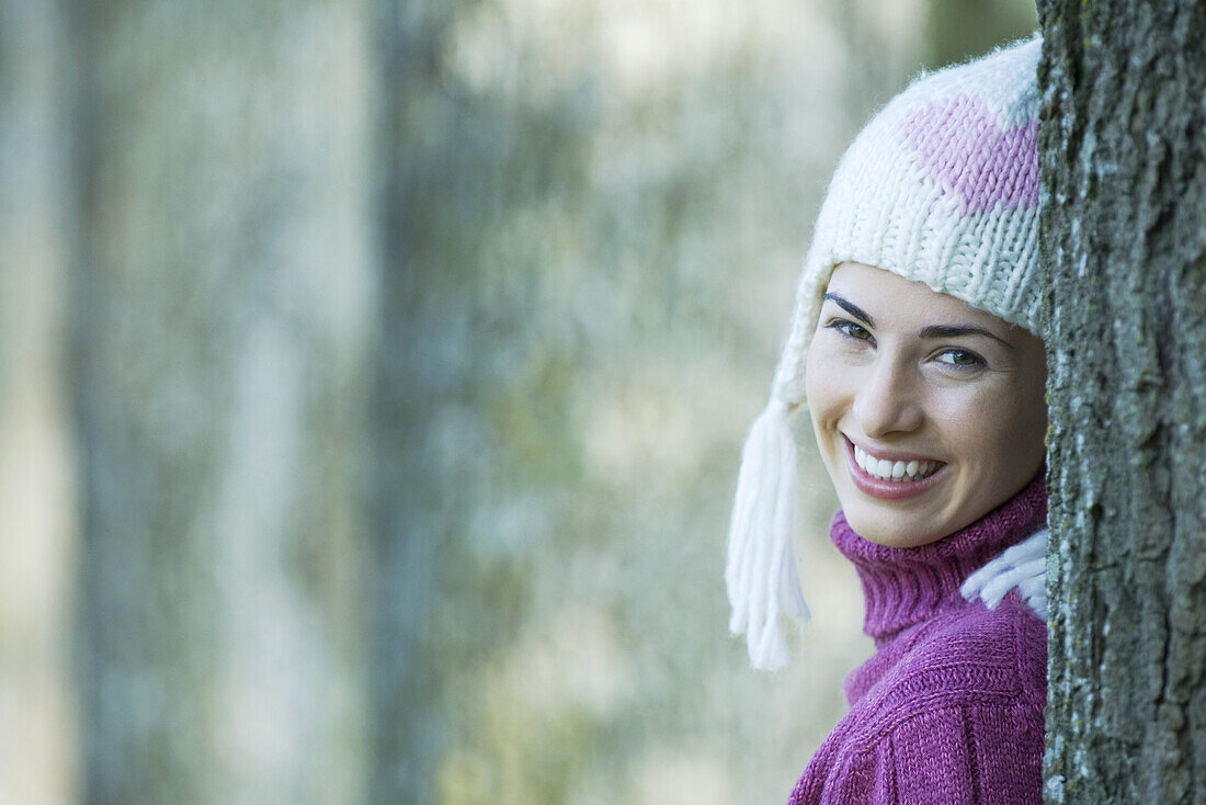 Young woman wearing knit hat, leaning against tree, smiling over shoulder at camera