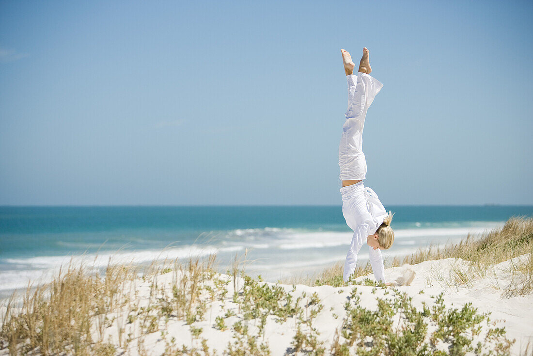Young woman doing handstand in dunes, ocean in background