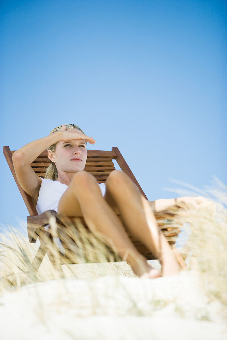 Young woman sitting in deck chair, in dunes, shading eyes with hand