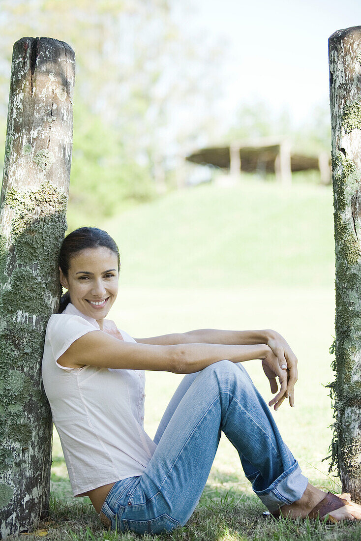 Woman sitting on grass, leaning against wooden post, full length
