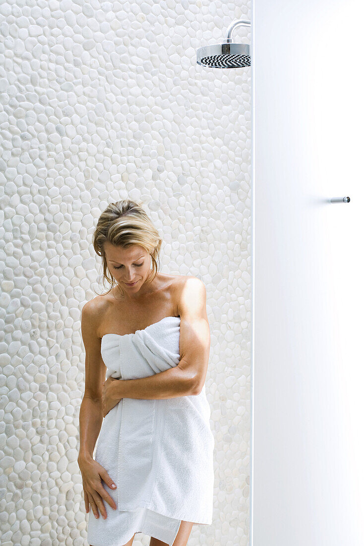 Frau steht unter der Dusche, in ein Handtuch gewickelt, blickt nach unten