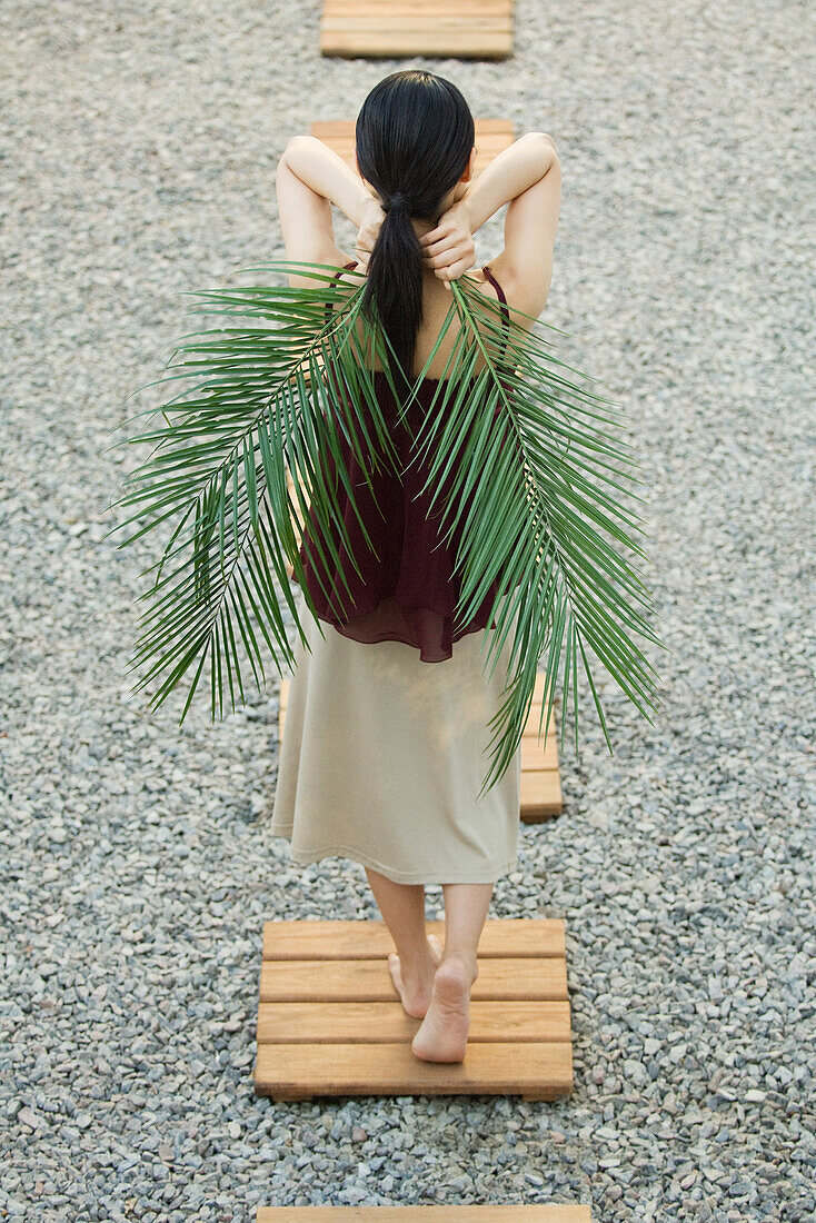 Woman walking on footpath, holding palm leaves behind back