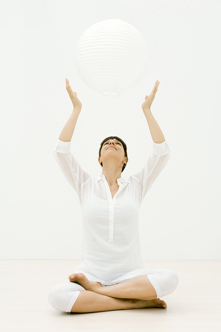 Woman sitting cross-legged on the ground with arms raised, looking up at sphere