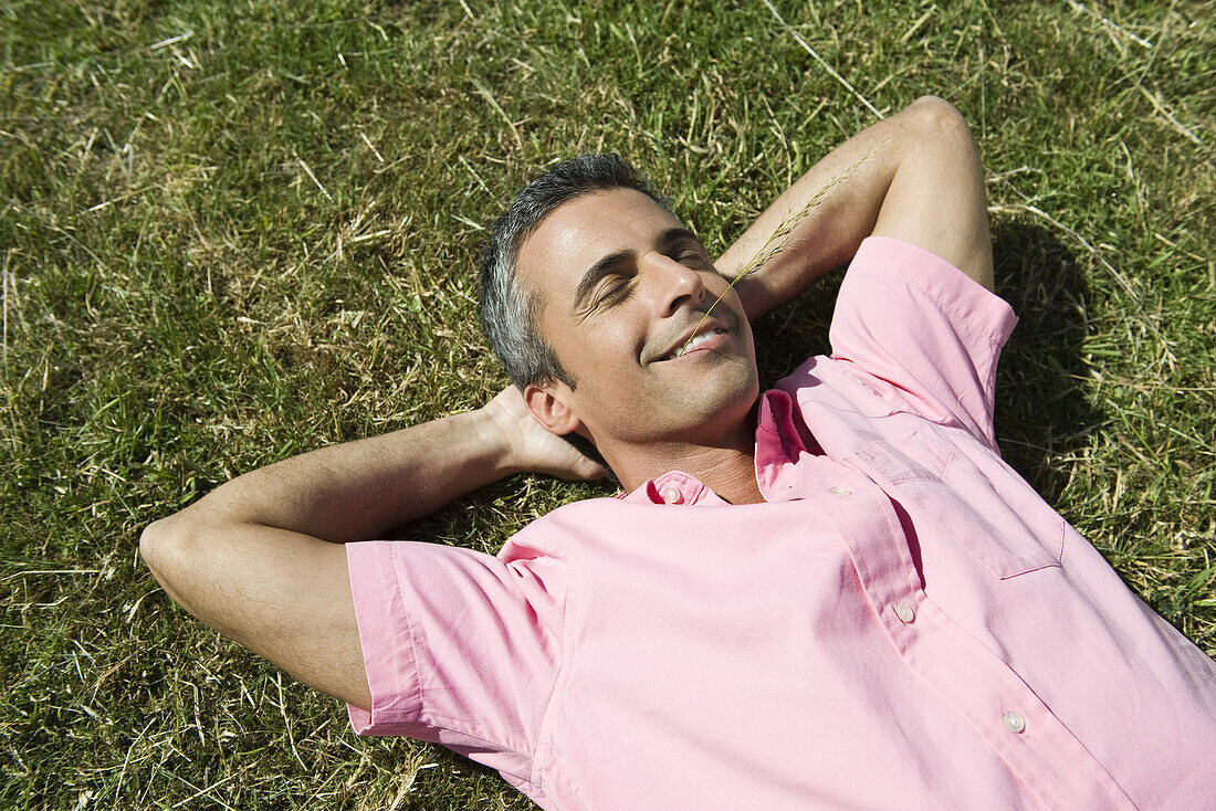 Man lying on grass with hands behind head and eyes closed, sprig of grass in mouth