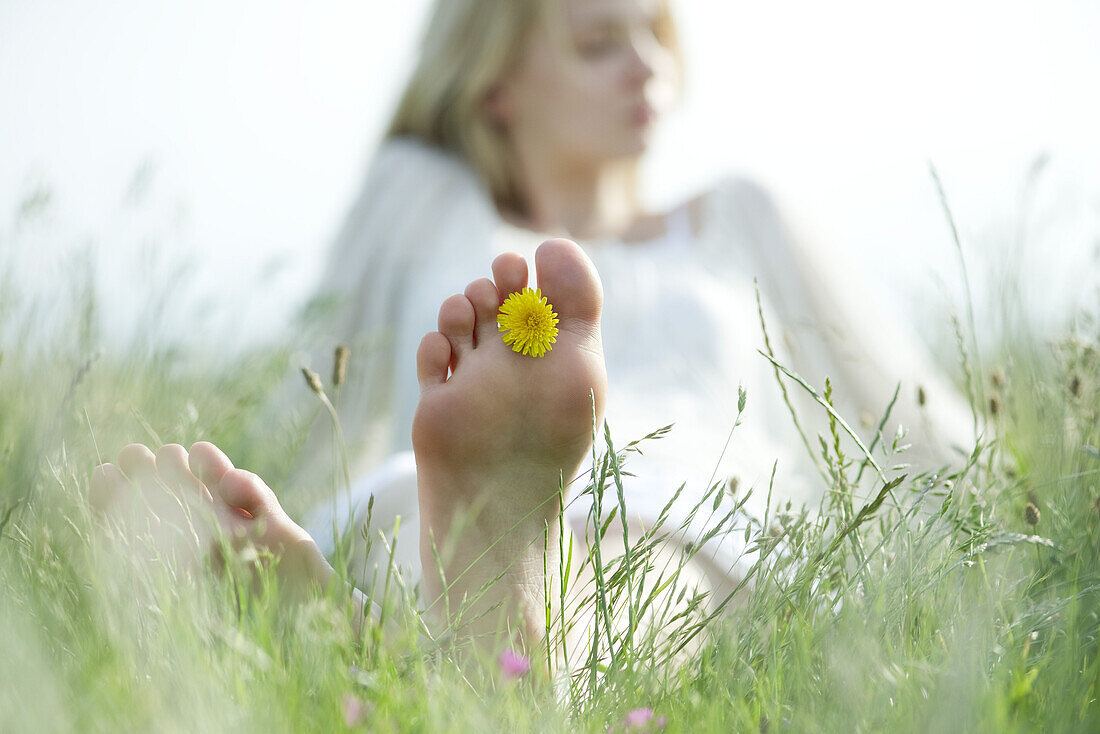 Barfuß sitzende junge Frau im Gras mit Löwenzahnblüten zwischen den Zehen, Ausschnitt