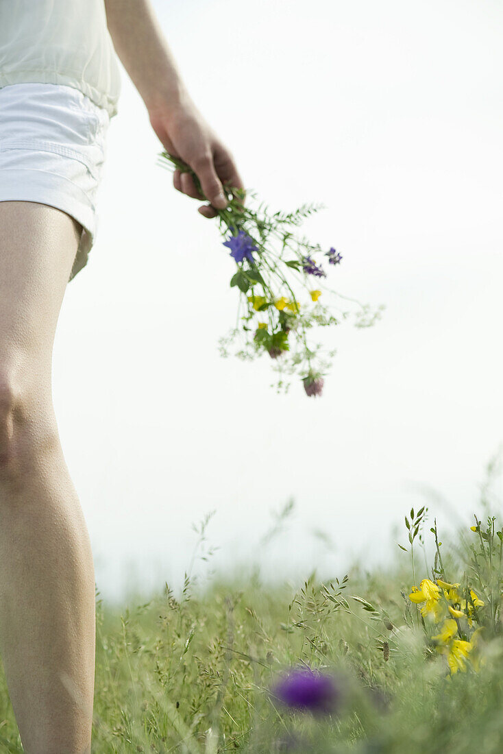 Young woman walking in meadow, carrying bouquet of wildflowers, cropped