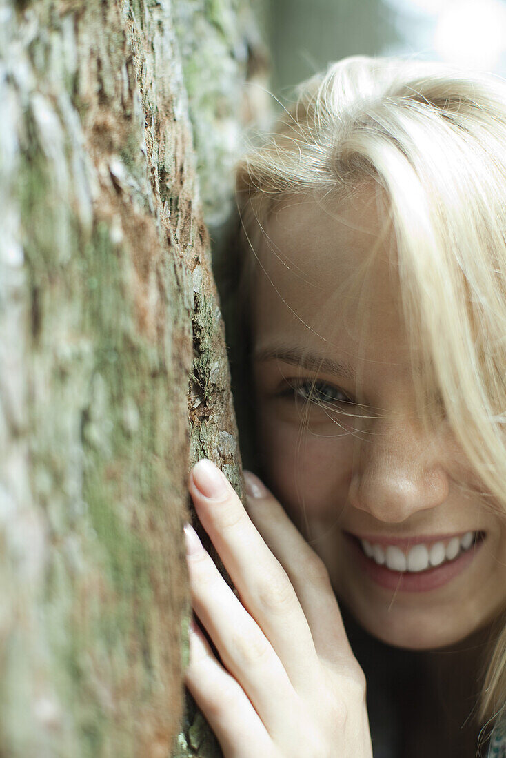 Smiling young woman leaning against tree trunk, cropped