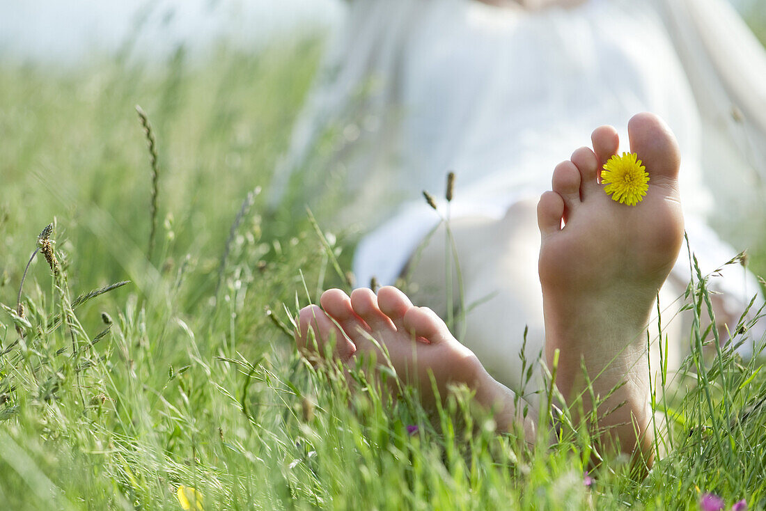 Barefoot woman sitting in grass, holding dandelion flower between toes