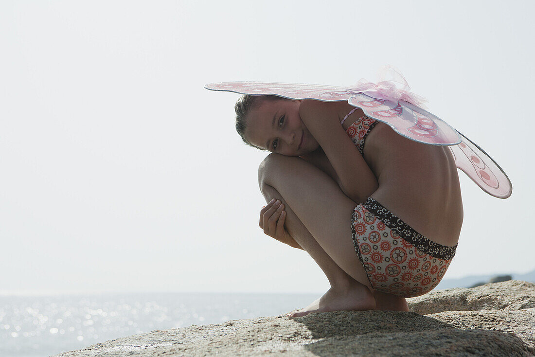 Girl in bikini wearing butterfly costume, crouching on rock