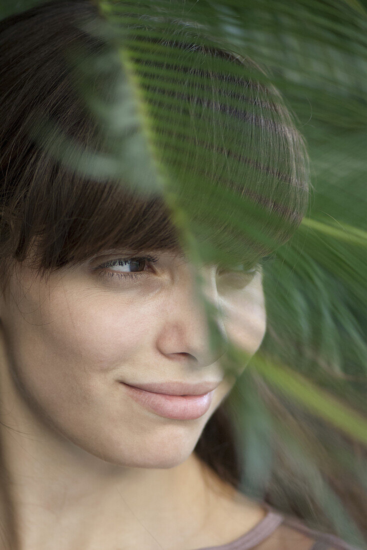 Young woman behind palm frond, looking away in thought